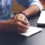 Manos anónimas tecleando en un portátil frente a siluetas de servidores en una oficina luminosa