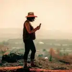 Manos sosteniendo un iPhone sobre una mochila de senderismo con cielo amplio al atardecer