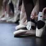 Manos ajustando cintas y volantes sobre telas de satén y gasa, con zapatillas de punta desenfocadas